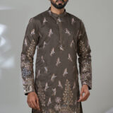 Man wearing a brown kurta with detailed bird and floral embroidery, paired with white trousers and black shoes, standing in a studio setting.