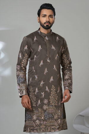Man wearing a brown kurta with detailed bird and floral embroidery, paired with white trousers and black shoes, standing in a studio setting.