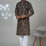 Man wearing a brown kurta with detailed bird and floral embroidery, paired with white trousers and black shoes, standing in a studio setting.