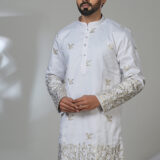 Man wearing a white kurta adorned with intricate gold floral embroidery on sleeves and hem, paired with white trousers and black shoes, standing in a studio setting.