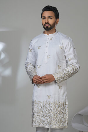 Man wearing a white kurta adorned with intricate gold floral embroidery on sleeves and hem, paired with white trousers and black shoes, standing in a studio setting.