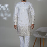 Man wearing a white kurta adorned with intricate gold floral embroidery on sleeves and hem, paired with white trousers and black shoes, standing in a studio setting.