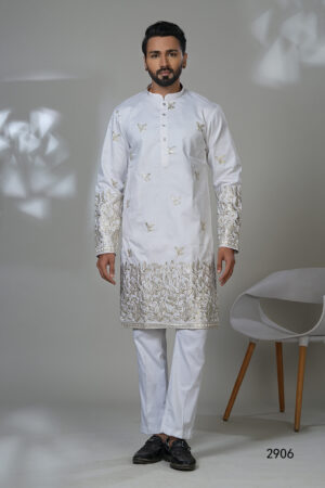 Man wearing a white kurta adorned with intricate gold floral embroidery on sleeves and hem, paired with white trousers and black shoes, standing in a studio setting.