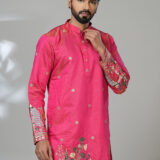 Man wearing a vibrant pink embroidered kurta with intricate floral and peacock designs, paired with white trousers and brown leather shoes, standing against a neutral background.