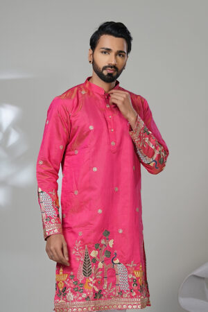 Man wearing a vibrant pink embroidered kurta with intricate floral and peacock designs, paired with white trousers and brown leather shoes, standing against a neutral background.