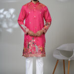 Man wearing a vibrant pink embroidered kurta with intricate floral and peacock designs, paired with white trousers and brown leather shoes, standing against a neutral background.