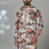 Man wearing a multicolor floral printed kurta with mirror work embroidery on the neckline, paired with white trousers and black loafers, standing indoors against a neutral backdrop.