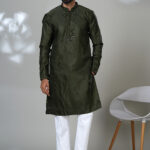 Man wearing a deep olive green embroidered kurta with tonal detailing and a band collar, paired with white trousers and black loafers, standing indoors against a neutral backdrop.