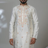 Man wearing an off-white kurta with peach-toned embroidery along the chest and small floral motifs, paired with white trousers and black loafers, standing indoors in a minimalist setting.
