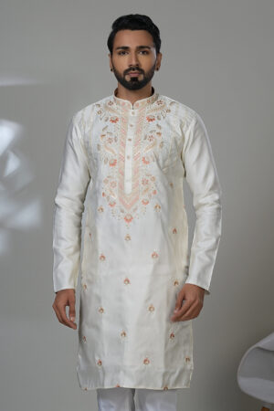 Man wearing an off-white kurta with peach-toned embroidery along the chest and small floral motifs, paired with white trousers and black loafers, standing indoors in a minimalist setting.