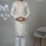 Man wearing an off-white kurta with peach-toned embroidery along the chest and small floral motifs, paired with white trousers and black loafers, standing indoors in a minimalist setting.