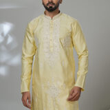 Man wearing a light yellow embroidered kurta with white thread and mirror detailing, paired with white trousers and black loafers, standing in a modern indoor studio setting.