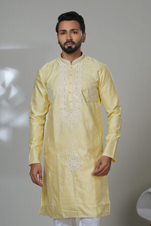 Man wearing a light yellow embroidered kurta with white thread and mirror detailing, paired with white trousers and black loafers, standing in a modern indoor studio setting.