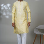 Man wearing a light yellow embroidered kurta with white thread and mirror detailing, paired with white trousers and black loafers, standing in a modern indoor studio setting.