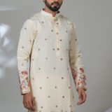 Man wearing an ivory embroidered kurta with pink and gold floral detailing on the sleeves and hem, paired with white churidar pants and brown loafers, standing in a modern indoor setting.
