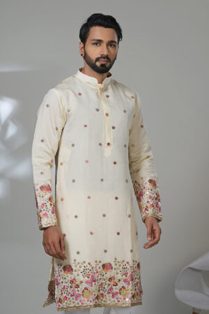 Man wearing an ivory embroidered kurta with pink and gold floral detailing on the sleeves and hem, paired with white churidar pants and brown loafers, standing in a modern indoor setting.