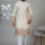 Man wearing an ivory embroidered kurta with pink and gold floral detailing on the sleeves and hem, paired with white churidar pants and brown loafers, standing in a modern indoor setting.