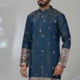 Man wearing a royal blue embroidered kurta with intricate multicolor detailing on the cuffs and hem, paired with white churidar pants and black loafers, standing in a modern indoor setting.