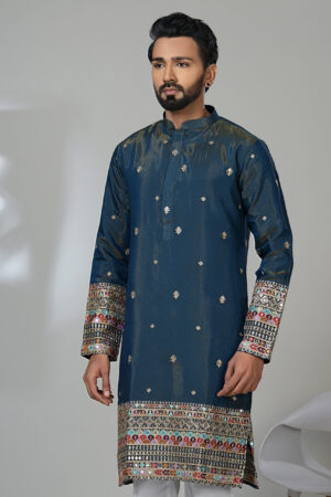 Man wearing a royal blue embroidered kurta with intricate multicolor detailing on the cuffs and hem, paired with white churidar pants and black loafers, standing in a modern indoor setting.