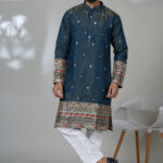 Man wearing a royal blue embroidered kurta with intricate multicolor detailing on the cuffs and hem, paired with white churidar pants and black loafers, standing in a modern indoor setting.