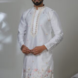 an in a white kurta with floral embroidery and mirror accents, worn with white trousers and black shoes.