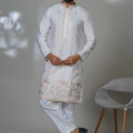 Man in a white kurta with floral embroidery and mirror accents, worn with white trousers and black shoes.