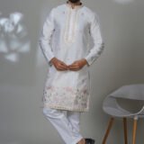 Man in a white kurta with floral embroidery and mirror accents, worn with white trousers and black shoes.