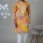 Man in a vibrant patchwork-printed kurta in shades of orange and yellow, styled with white pants and black loafers.