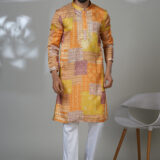 Man in a vibrant patchwork-printed kurta in shades of orange and yellow, styled with white pants and black loafers.