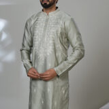 Man in a sage green embroidered kurta and white trousers, standing in a modern indoor setting next to a white chair.