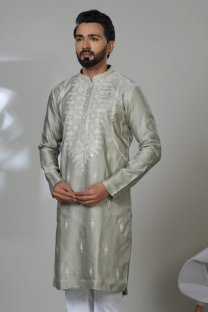 Man in a sage green embroidered kurta and white trousers, standing in a modern indoor setting next to a white chair.