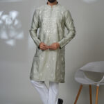 Man in a sage green embroidered kurta and white trousers, standing in a modern indoor setting next to a white chair.