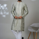 Man in a sage green embroidered kurta and white trousers, standing in a modern indoor setting next to a white chair.