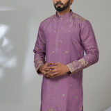 Man wearing a mauve kurta with gold ethnic embroidery and white trousers, posing in a minimalistic indoor setting.