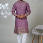 Man wearing a mauve kurta with gold ethnic embroidery and white trousers, posing in a minimalistic indoor setting.