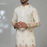 Man wearing a cream embroidered kurta with floral detailing and white trousers, standing next to a modern white chair.
