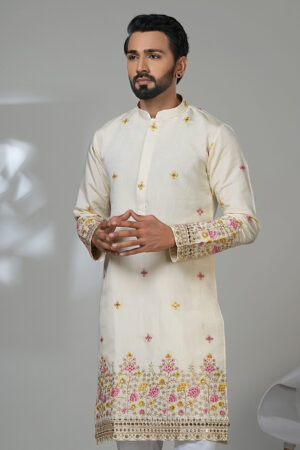 Man wearing a cream embroidered kurta with floral detailing and white trousers, standing next to a modern white chair.
