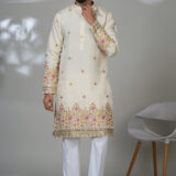 Man wearing a cream embroidered kurta with floral detailing and white trousers, standing next to a modern white chair.