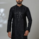 Man wearing a black kurta with mirror work and white trousers, posing in a studio with a modern chair in the background.