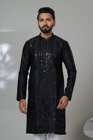 Man wearing a black kurta with mirror work and white trousers, posing in a studio with a modern chair in the background.