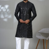 Man wearing a black kurta with mirror work and white trousers, posing in a studio with a modern chair in the background.