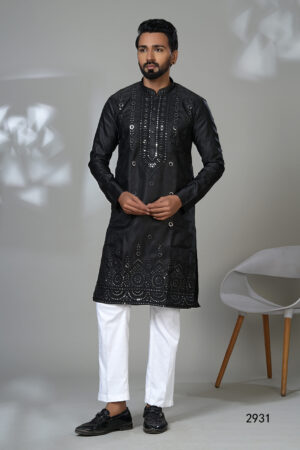 Man wearing a black kurta with mirror work and white trousers, posing in a studio with a modern chair in the background.