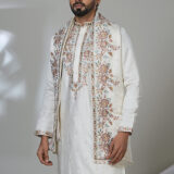 Man wearing an ivory embroidered kurta with floral dupatta and jacket, white trousers, and brown loafers, standing in a modern setting.