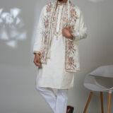 Man wearing an ivory embroidered kurta with floral dupatta and jacket, white trousers, and brown loafers, standing in a modern setting.