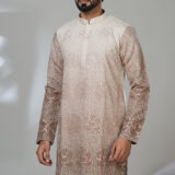 Man wearing a beige and brown embroidered kurta with white trousers and black shoes, standing in a minimalistic room with a white chair.