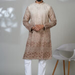 Man wearing a beige and brown embroidered kurta with white trousers and black shoes, standing in a minimalistic room with a white chair