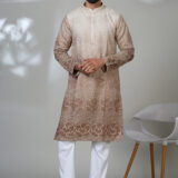 Man wearing a beige and brown embroidered kurta with white trousers and black shoes, standing in a minimalistic room with a white chair