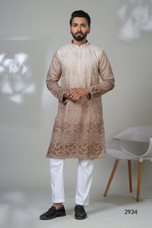 Man wearing a beige and brown embroidered kurta with white trousers and black shoes, standing in a minimalistic room with a white chair