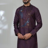 Man wearing a plum-colored embroidered kurta with blue peacock motifs, white pants, and brown formal loafers, standing beside a modern white chair
