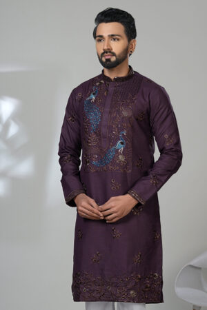 Man wearing a plum-colored embroidered kurta with blue peacock motifs, white pants, and brown formal loafers, standing beside a modern white chair